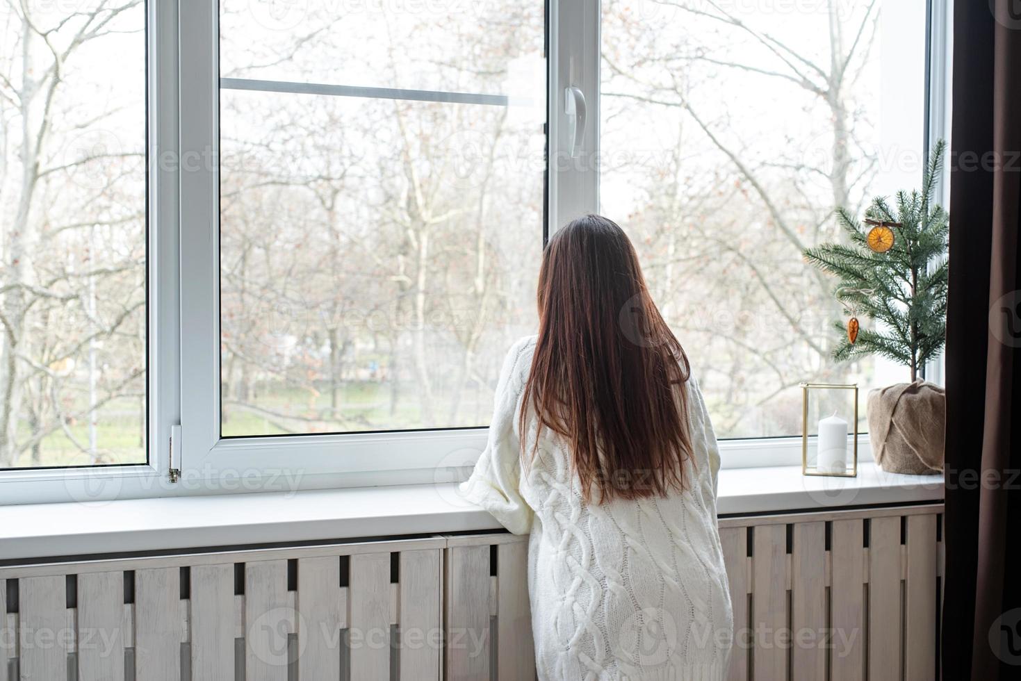 Woman in warm white winter sweater standing next to the window at home at christmas eve holding cup with marshmallows, fir tree behind photo
