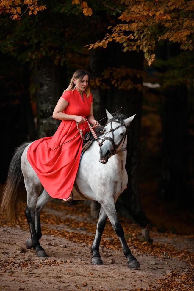 Riding a horse, walking in an autumn forest, a woman riding a horse in a long red dress with bare feet. photo