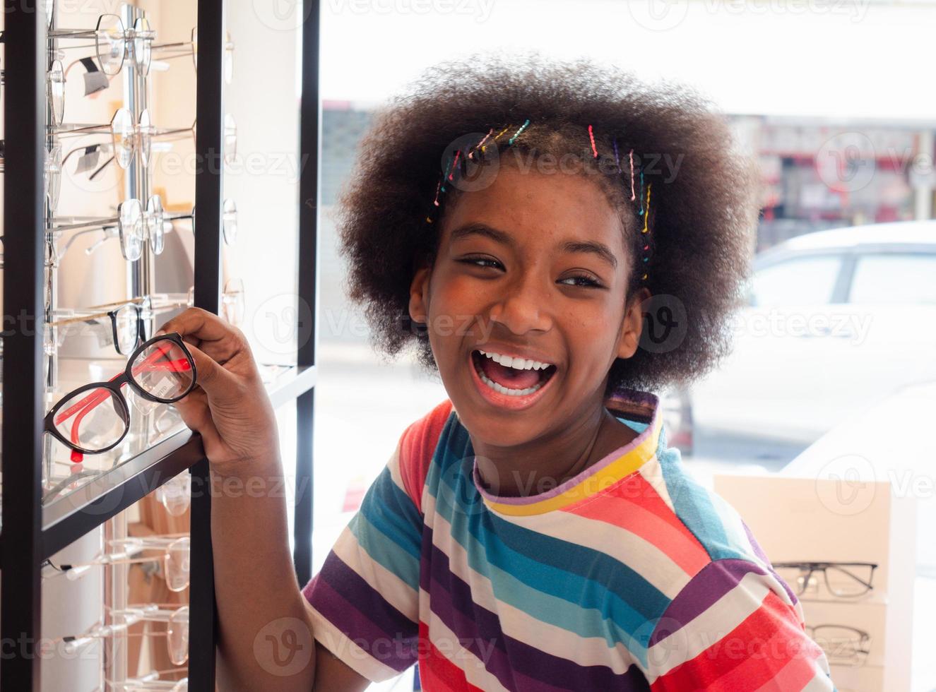 African girl in the optic store looking in the mirror for choosing a new eyeglasses frame. health care, medicine, people, eyesight and technology concept photo