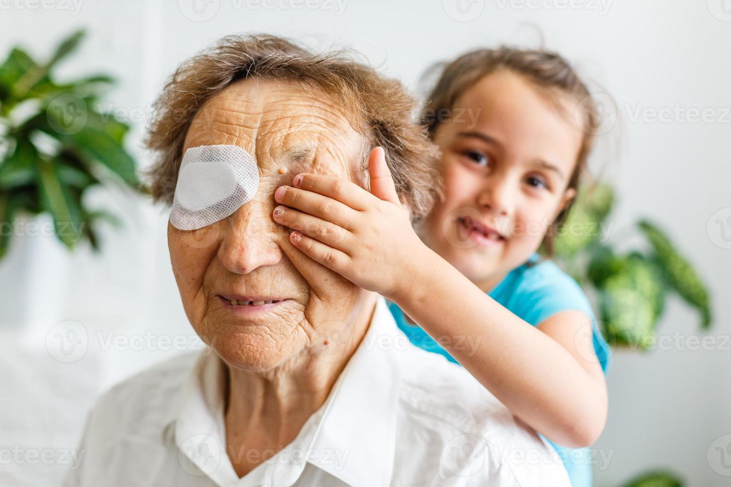 Little girl and senior woman at home celebration together sitting on sofa granddaughter covering eyes of granny making surprise smiling joyful photo