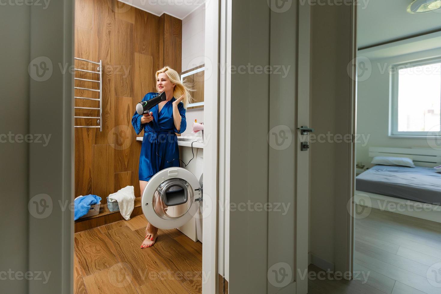 Beautiful girl using a hair dryer and smiling. Smiling woman drying hair with hair dry machine. Happy girl looking at mirror while using dryer in the bathroom. photo