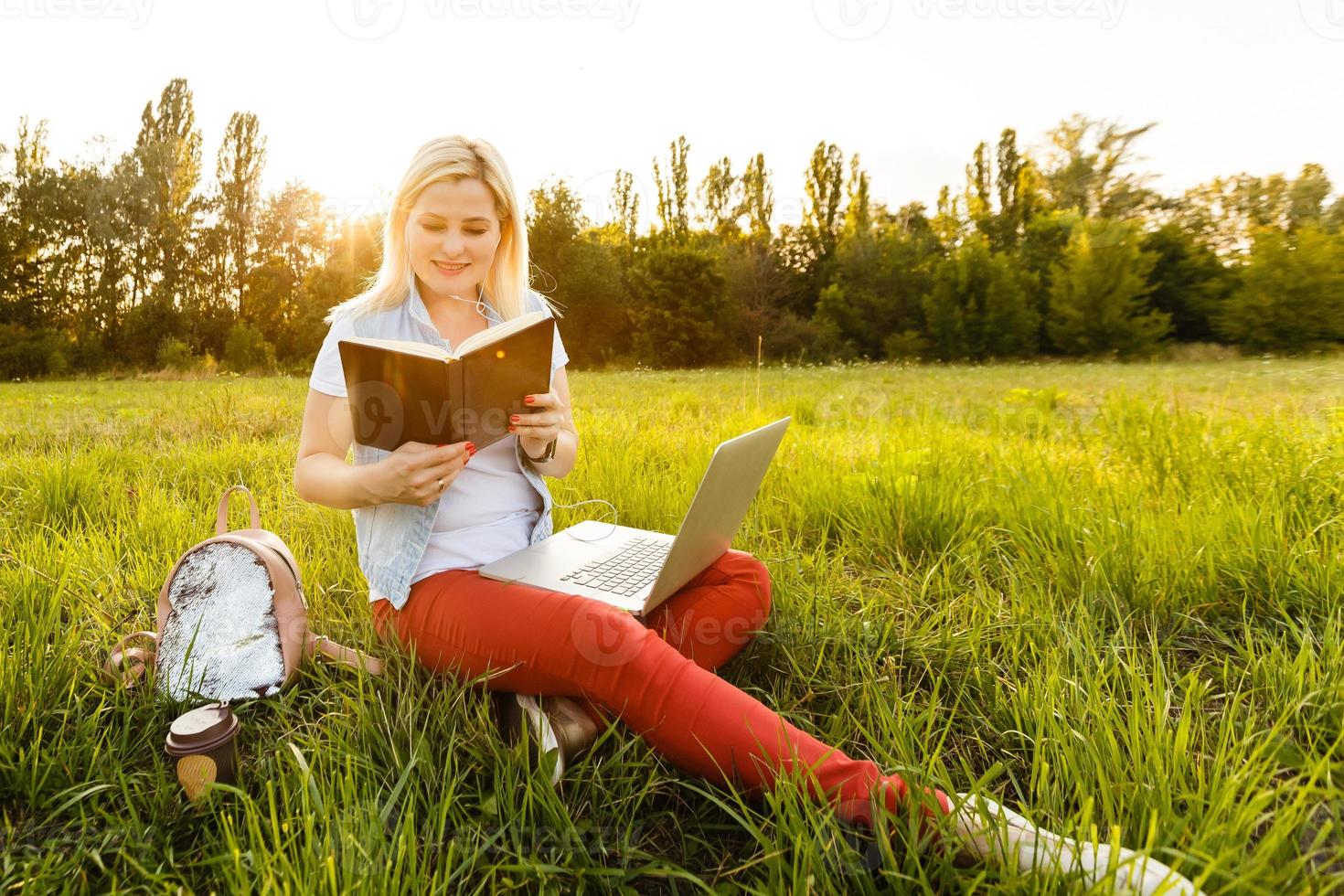 Young woman with laptop sits on the grass in the park on a sunny day. Laughing blonde in a white tank top and shorts. Online training, remote work and communication in social networks photo