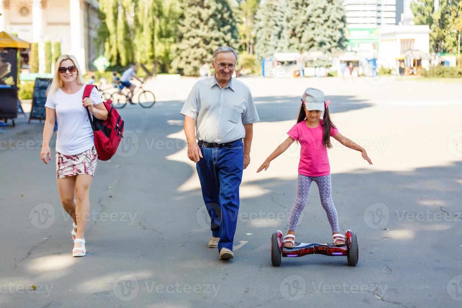 Little smiling girl learning to ride a hoverboard with her parents outdoors photo