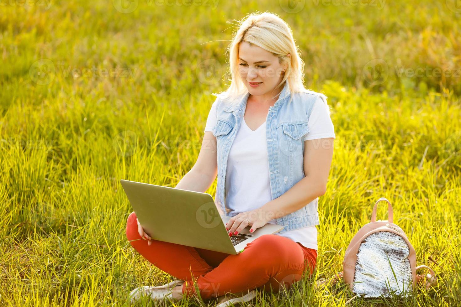 Young woman with laptop sits on the grass in the park on a sunny day. Laughing blonde in a white tank top and shorts. Online training, remote work and communication in social networks photo