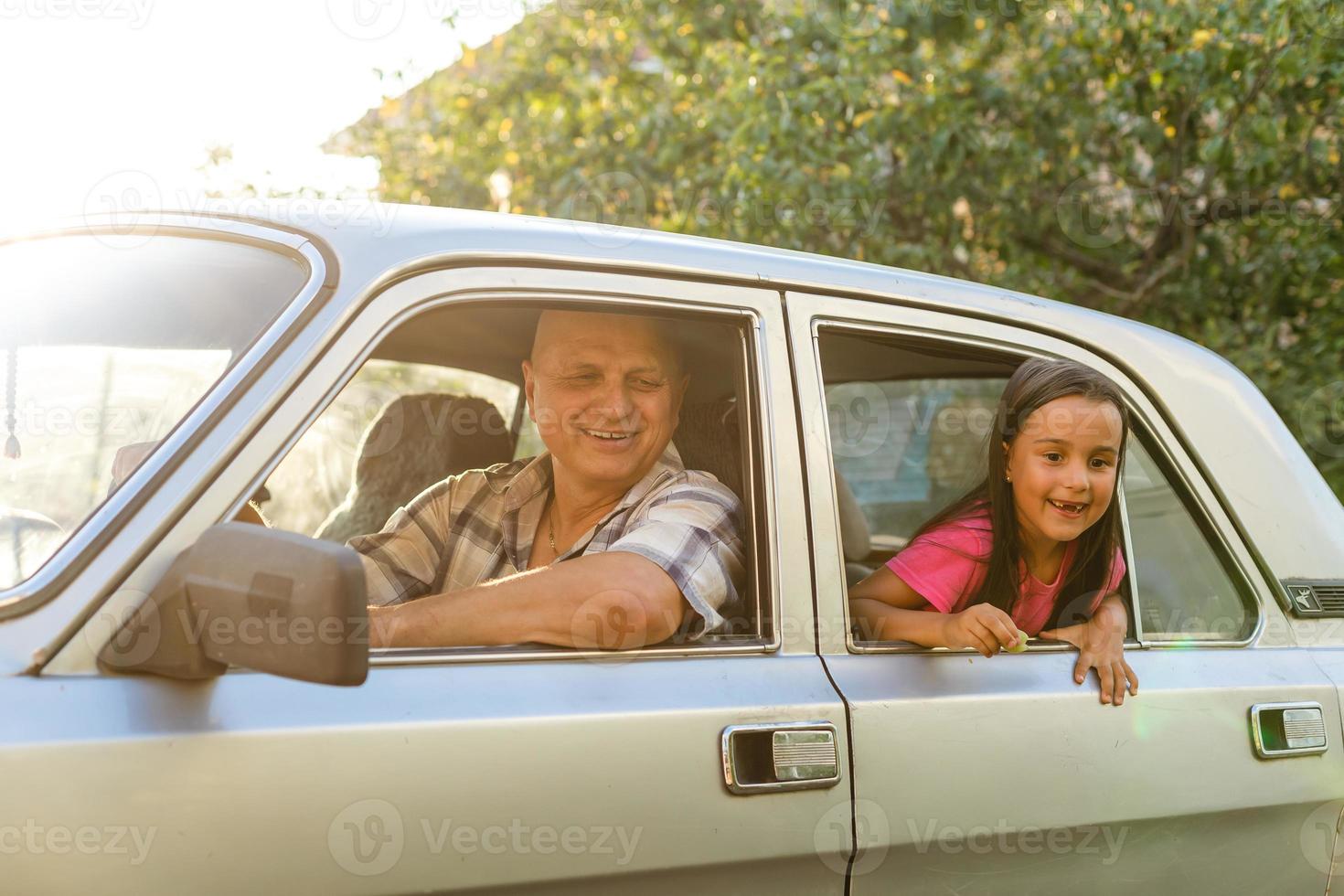 Senior man driving car with his granddaughter sitting behind with dog, looking through window photo