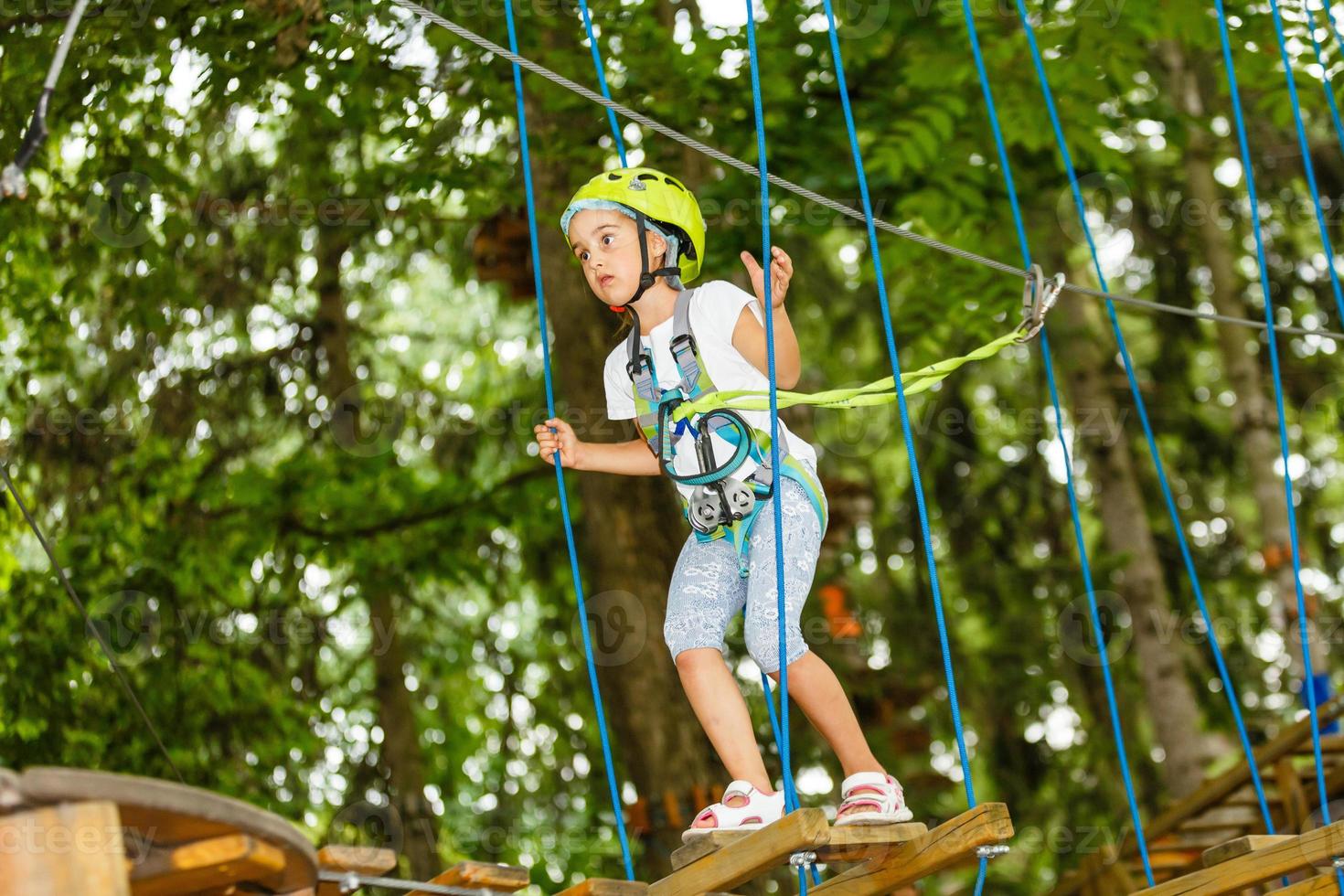 Portrait of little smiling girl in helmet and harness on trail in sky rope park in summer. photo