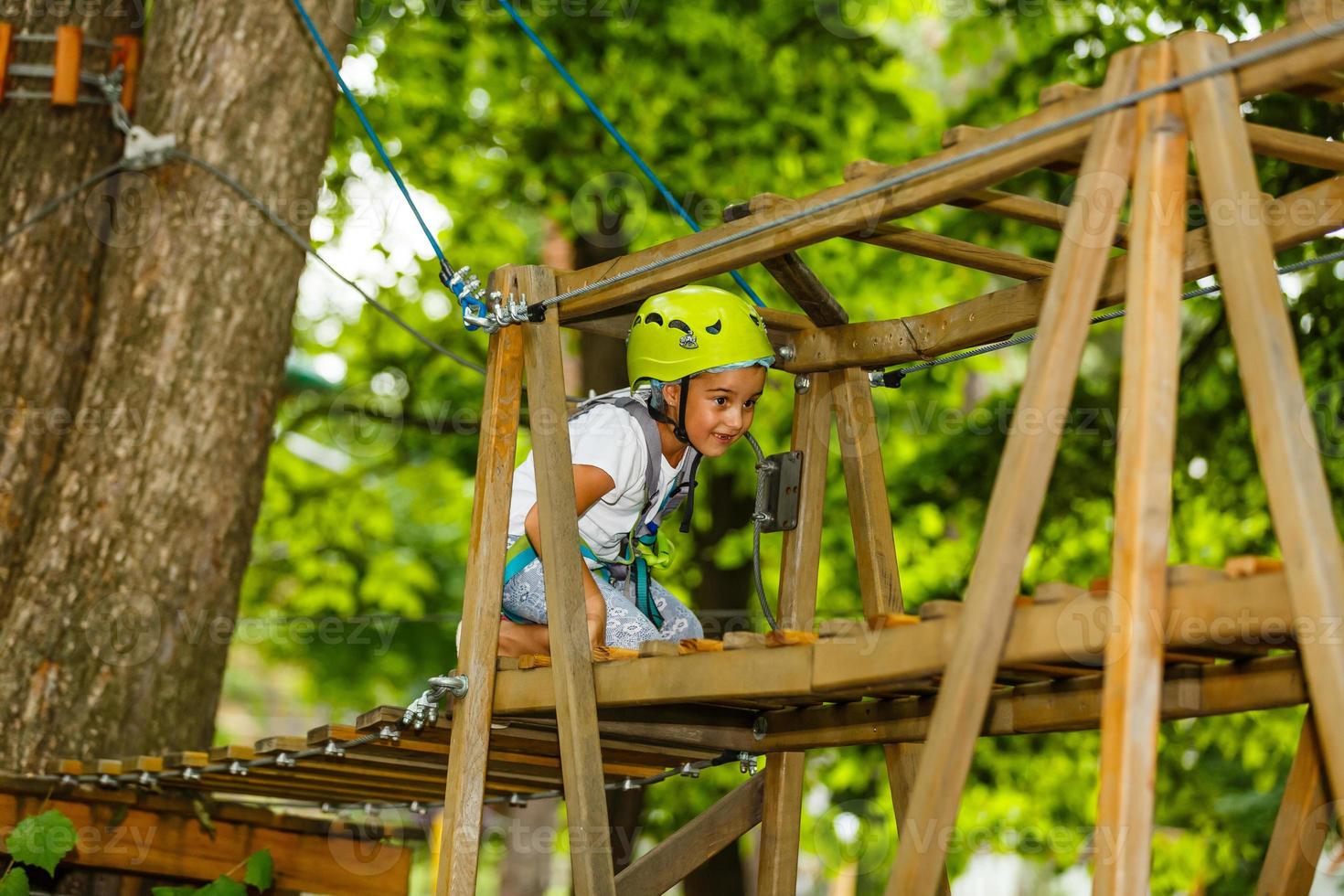Portrait of little smiling girl in helmet and harness on trail in sky rope park in summer. photo