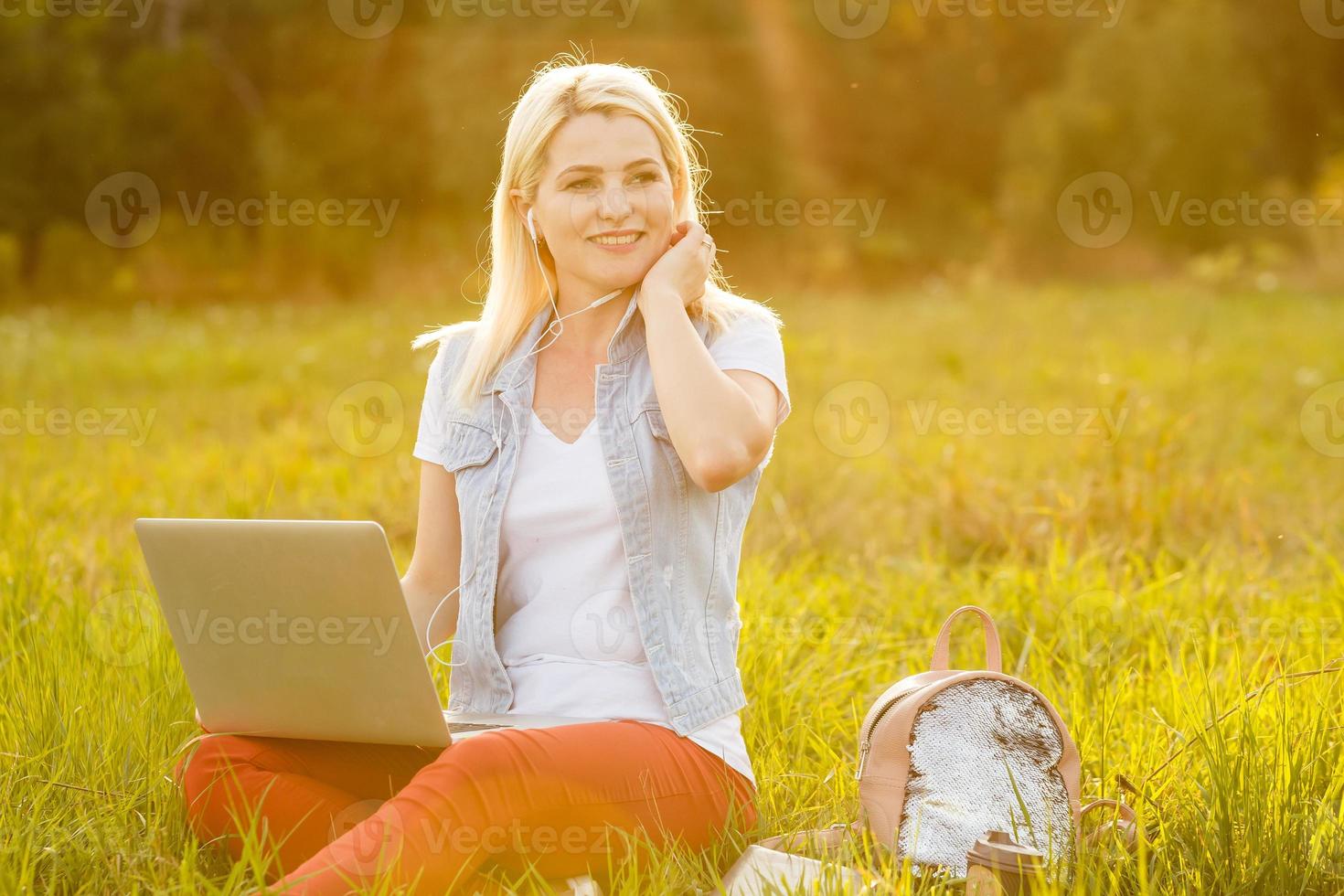 Young woman with laptop sits on the grass in the park on a sunny day. Laughing blonde in a white tank top and shorts. Online training, remote work and communication in social networks photo