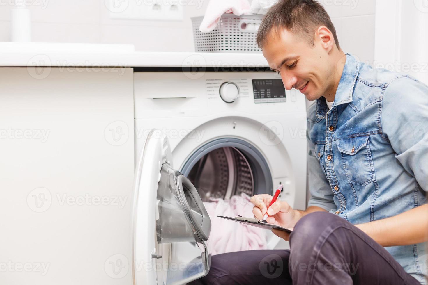 cropped shot of handyman with toolbelt and clipboard checking washing machine in bathroom photo