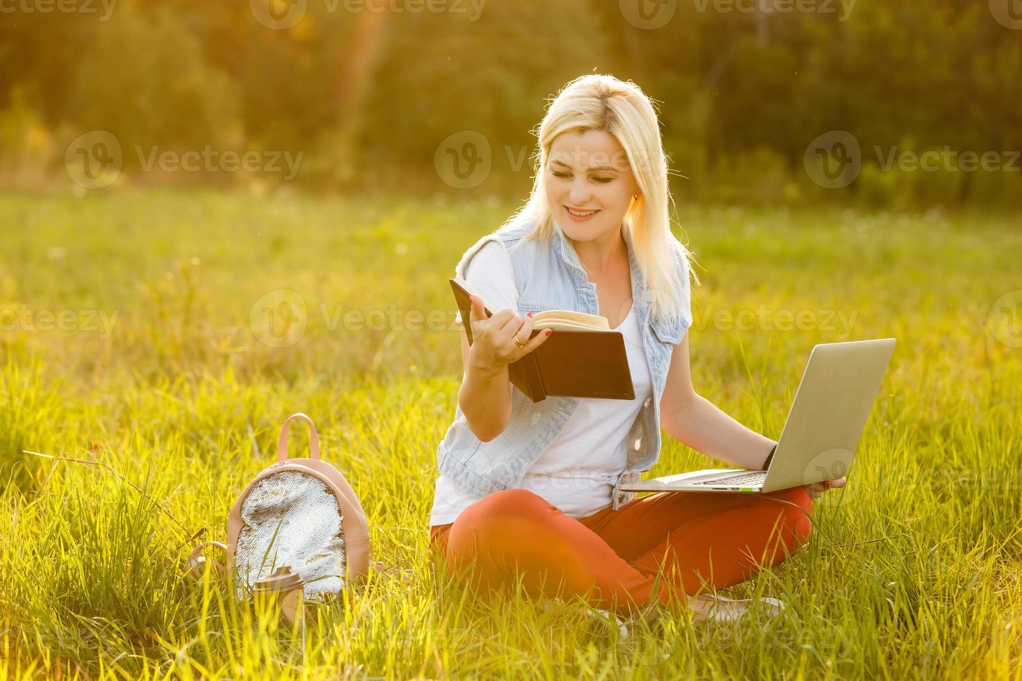 Portrait of pretty young woman sitting on green grass in park with legs crossed during summer day while using laptop for video call photo