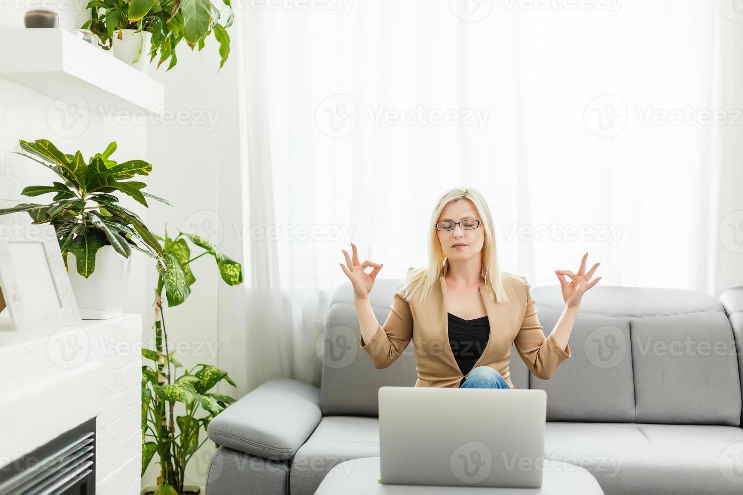 mujer joven meditando sentada frente a la computadora portátil, tomando una pausa, oficina ocupada y estresante, cura para la sobrecarga de trabajo, meditación de un momento, adorando la computadora portátil foto