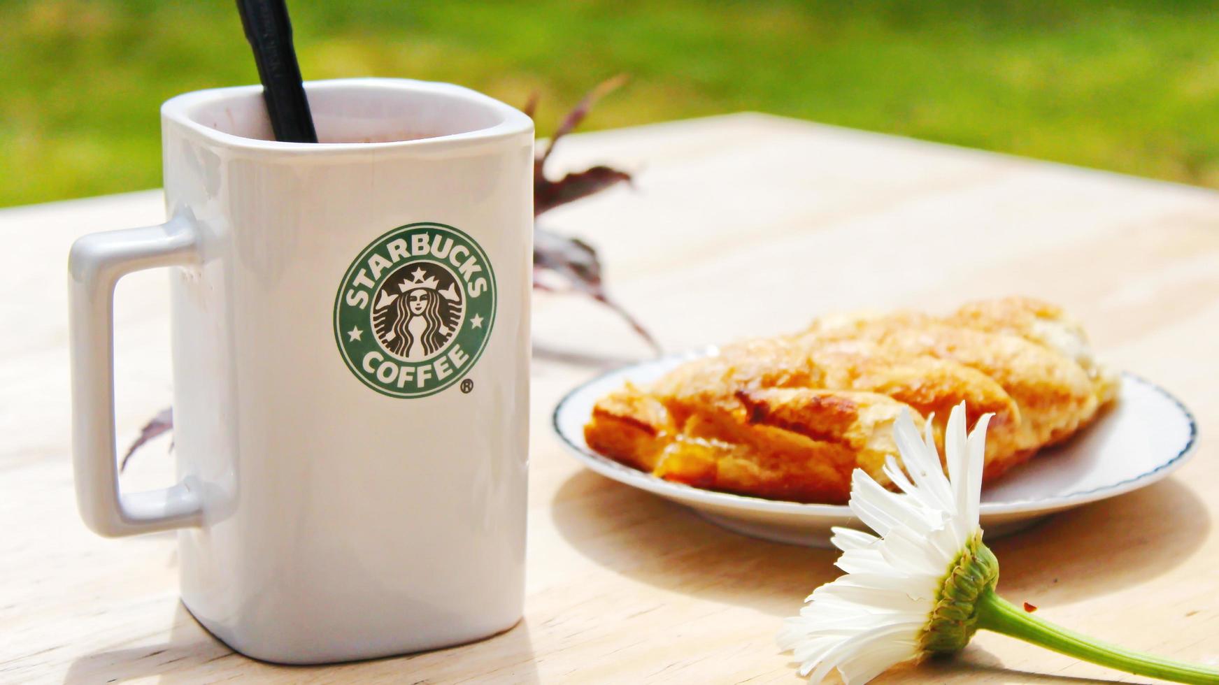 WASHINGTON, USA - August 01 2022  Coffee mug with Starbucks logo on front, white bakery on plate. Place it on a wooden table in the garden where the morning sun shines. photo