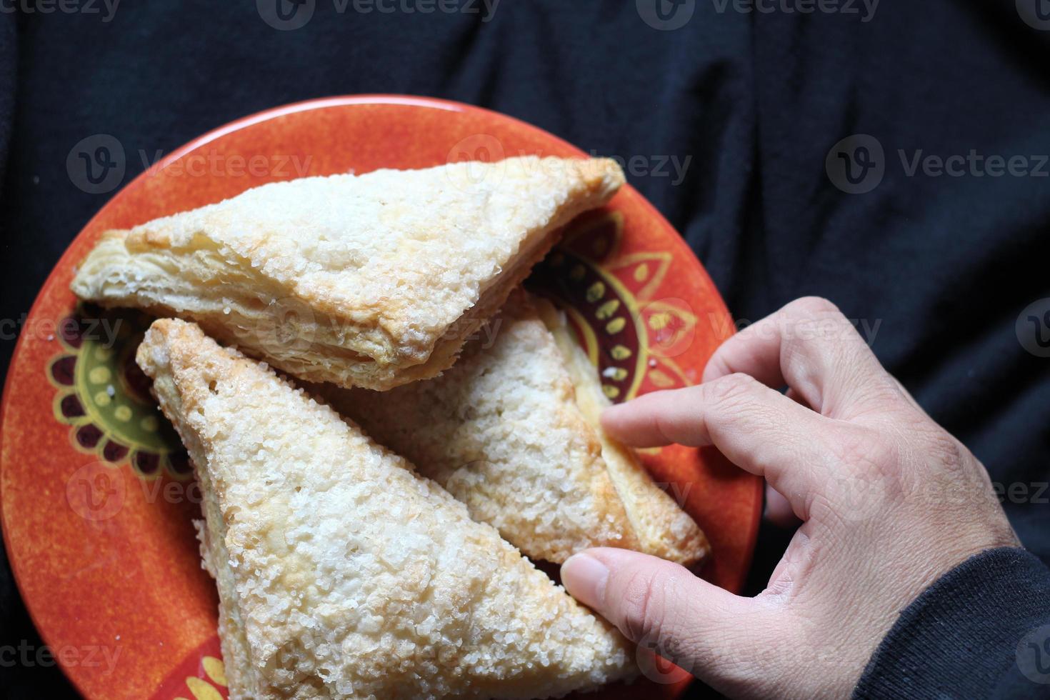 Man's hand holding Turnovers Apple, Apple Pie, Served in an Orange Plate on black cloth background, Turnovers Apple is a popular dish in America - Flat-Lay photo