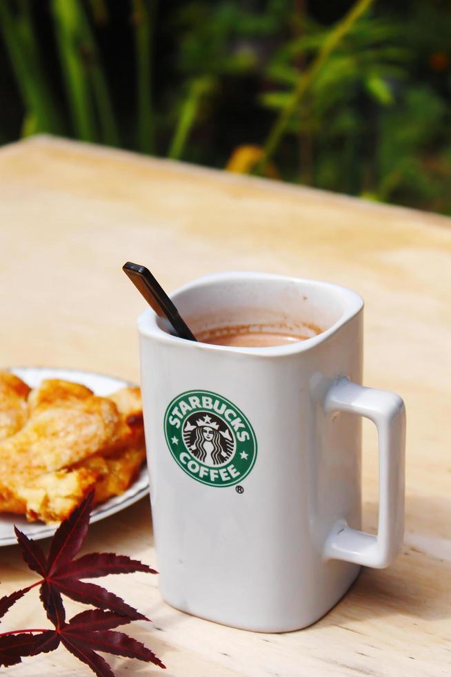 WASHINGTON, USA - August 01 2022  Coffee mug with Starbucks logo on front, white bakery on plate. Place it on a wooden table in the garden where the morning sun shines. photo