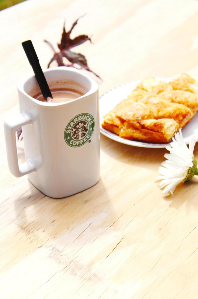 WASHINGTON, USA - August 01 2022  Coffee mug with Starbucks logo on front, white bakery on plate. Place it on a wooden table in the garden where the morning sun shines. photo