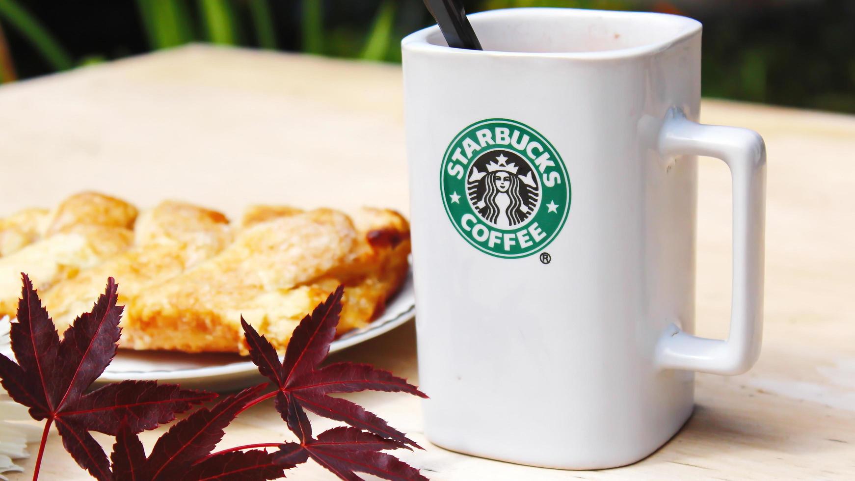 WASHINGTON, USA - August 01 2022  Coffee mug with Starbucks logo on front, white bakery on plate. Place it on a wooden table in the garden where the morning sun shines. photo