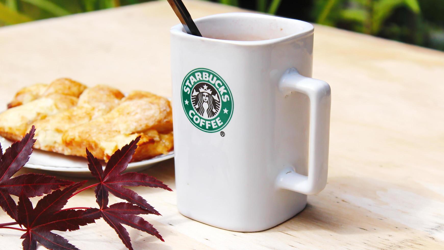 WASHINGTON, USA - August 01 2022  Coffee mug with Starbucks logo on front, white bakery on plate. Place it on a wooden table in the garden where the morning sun shines. photo