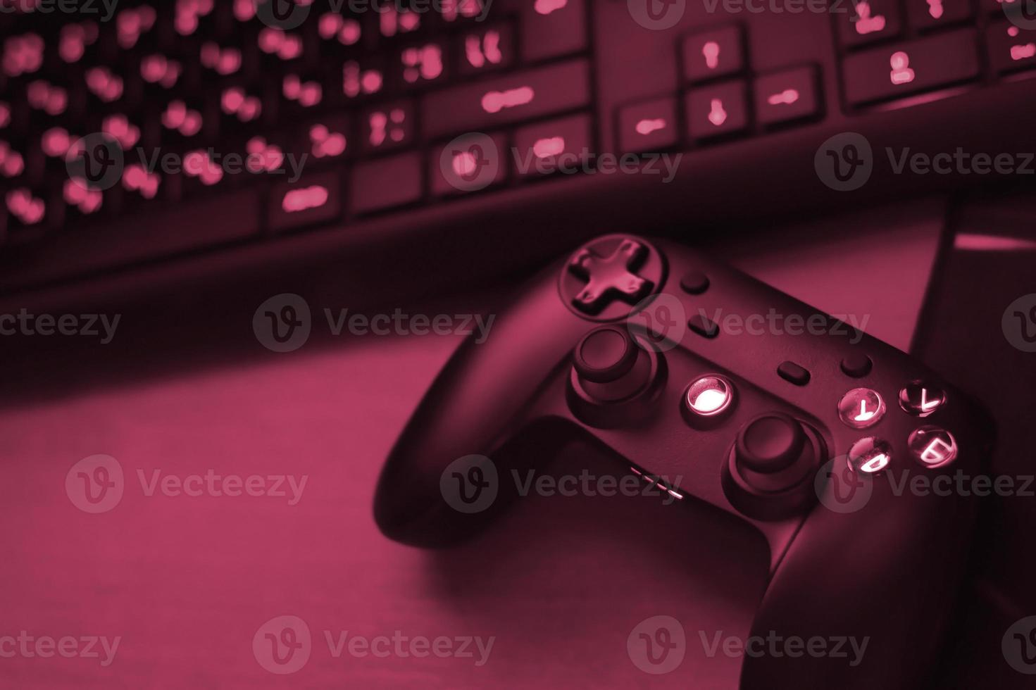 Modern gamepad lies with pc keyboard on table in dark playroom scene. Gameplay streaming and video game walkthroughts concept Image toned in Viva Magenta, color of the 2023 year photo