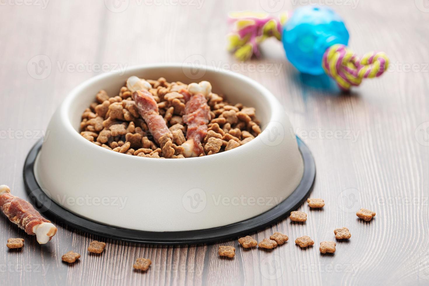 un tazón con comida para perros, golosinas para perros y juguetes en un piso de madera. foto