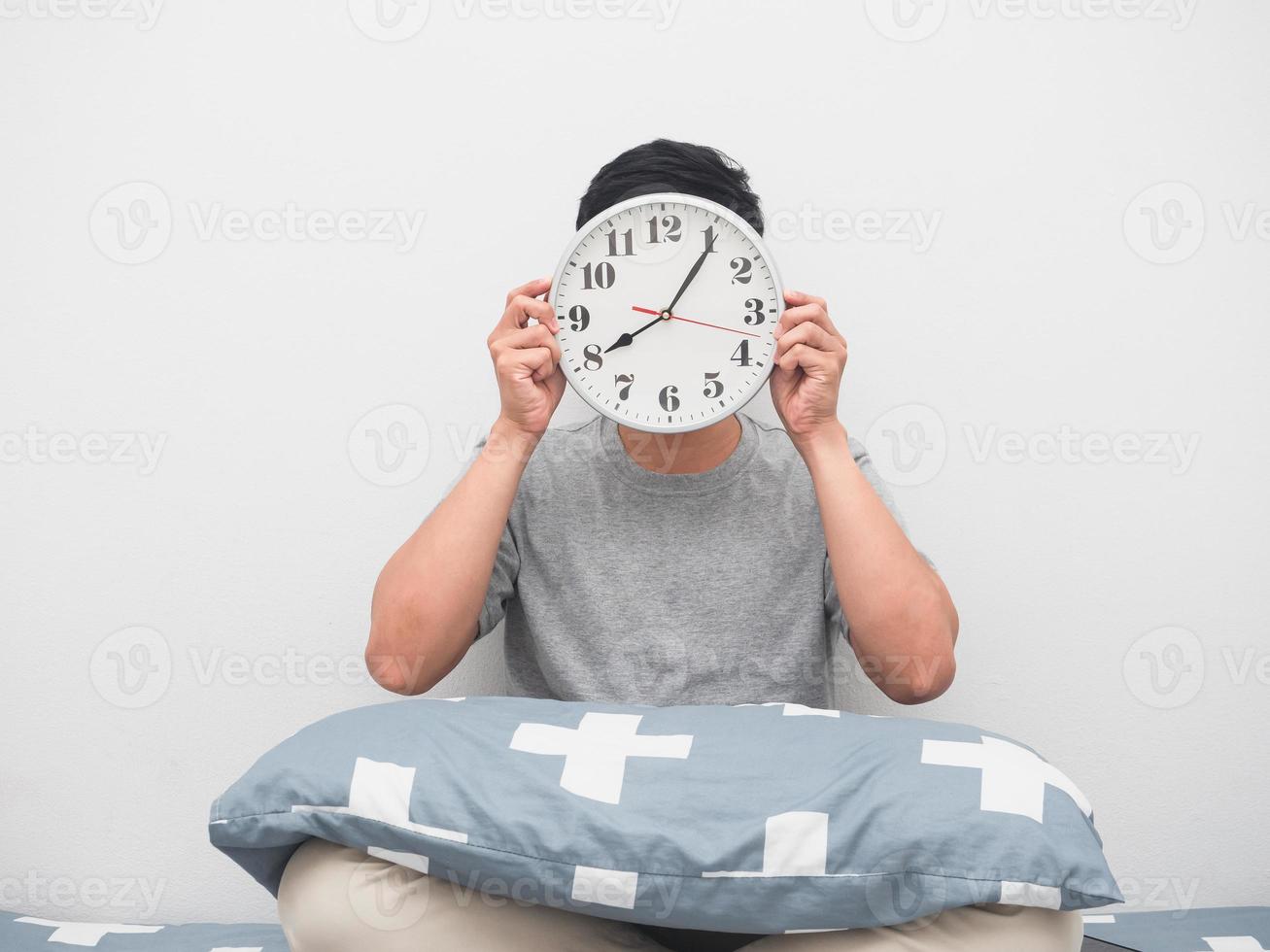 Man sit on the bed holding analog clock close his face,Wake up late concept photo