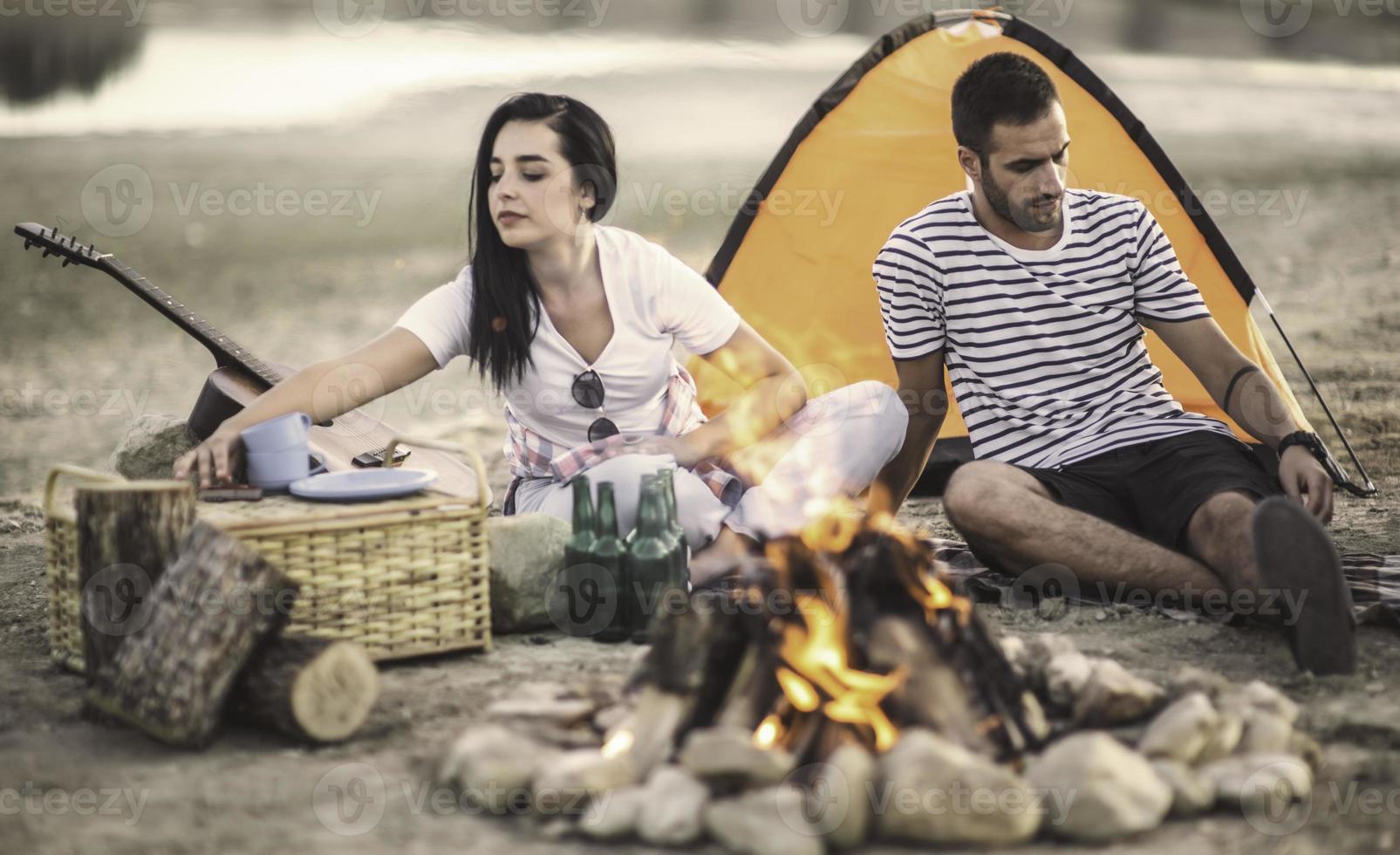 concepto de vacaciones de picnic. hermosa pareja divirtiéndose haciendo barbacoa en la hoguera y relajándose junto a un lago. foto