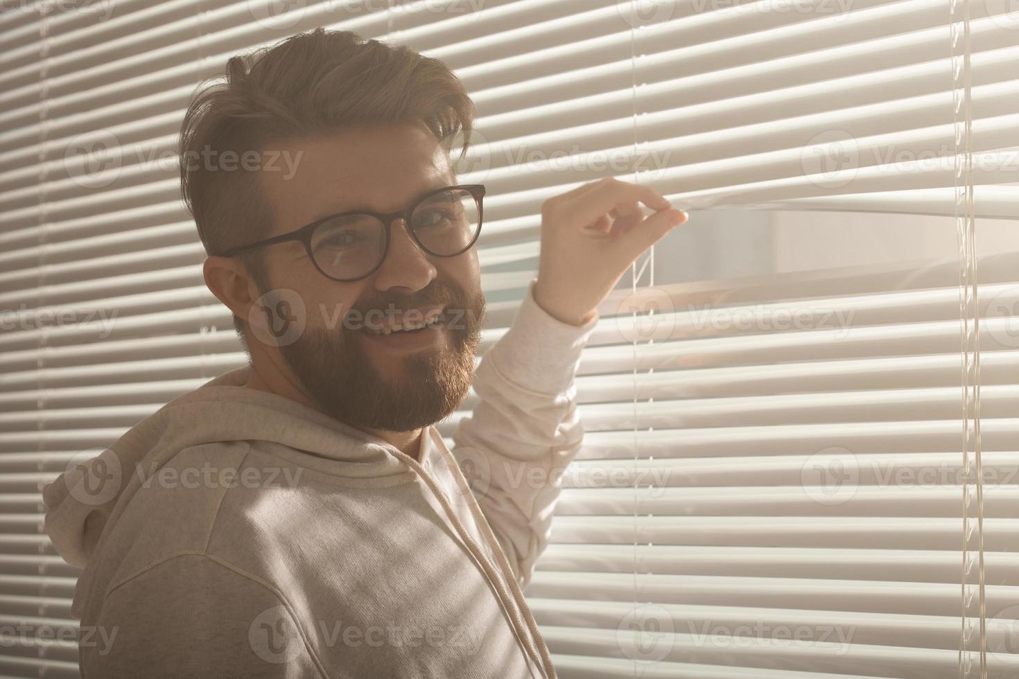 Young man with beard peeks through hole in the window blinds and looks out into the street. Surveillance and curiosity concept photo