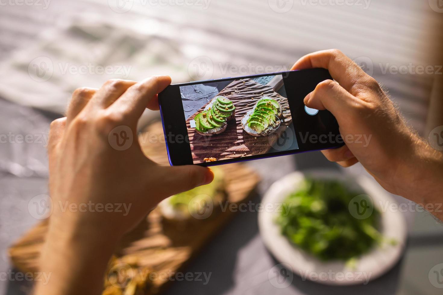 Hands take pictures on smartphone of two beautiful healthy sour cream and avocado sandwiches lying on board on the table. Social media and food concept photo