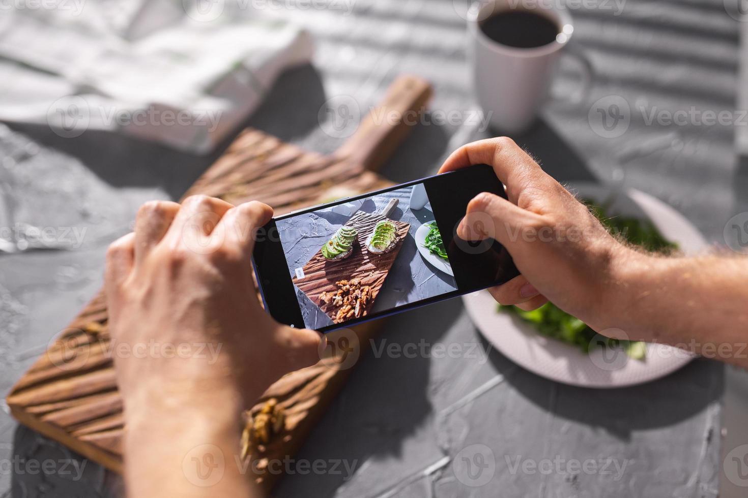 Hands take pictures on smartphone of two beautiful healthy sour cream and avocado sandwiches lying on board on the table. Social media and food concept photo