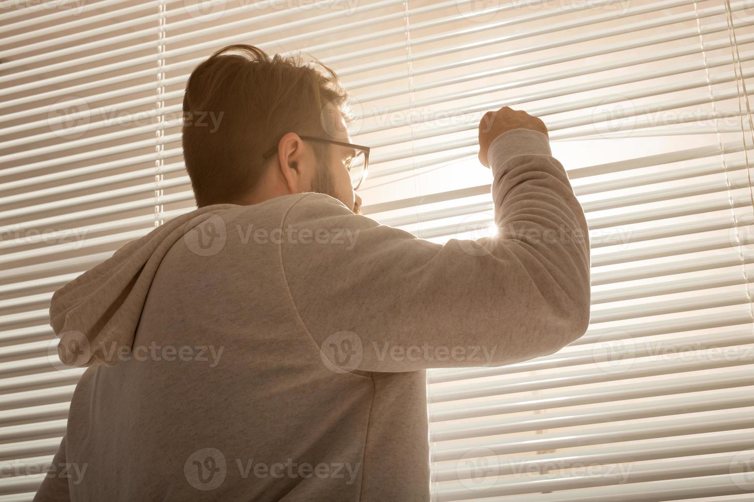 Rear view of young stylish man peeking through hole in window blinds and looking out into street. Concept of enjoying the morning sun and positivity photo