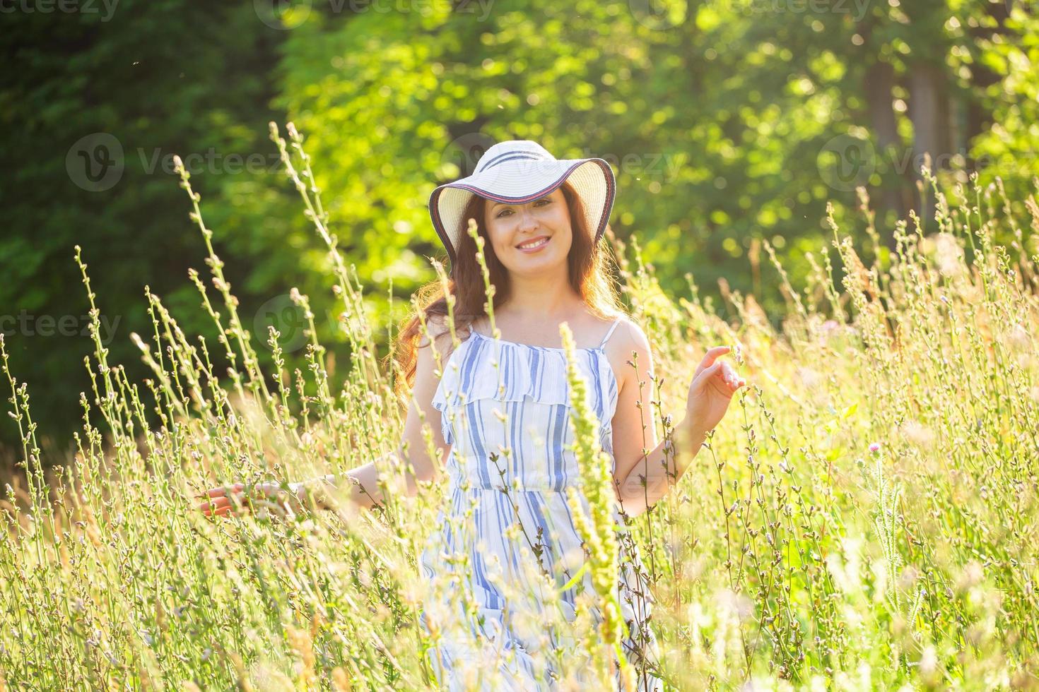 mujer joven feliz con el pelo largo con sombrero y vestido caminando por el bosque de verano en un día soleado. concepto de alegría de verano foto