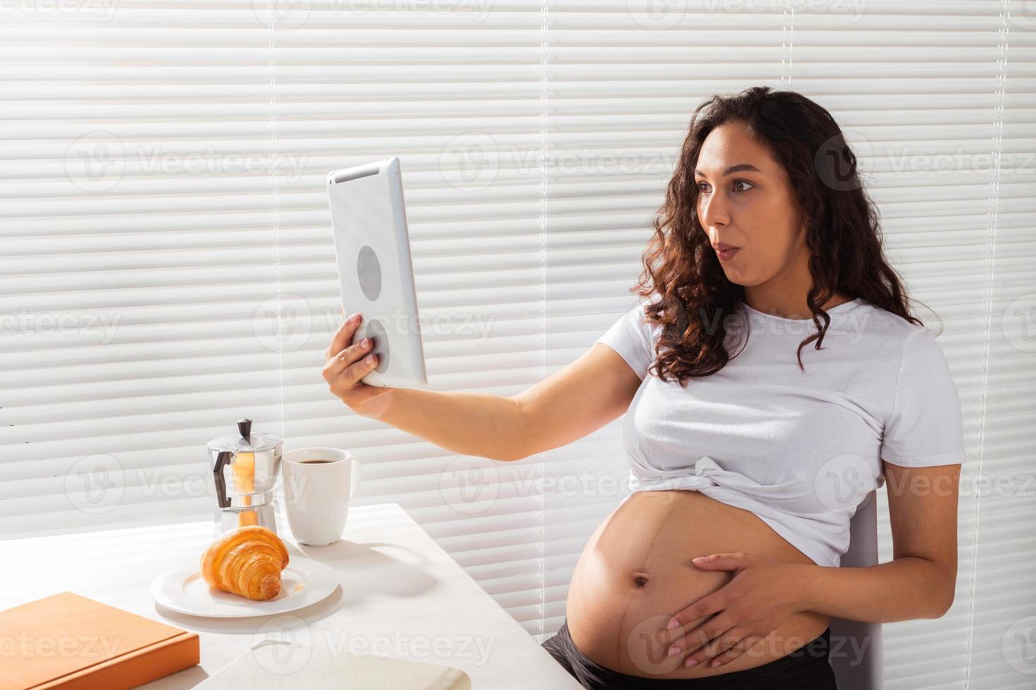 Happy pregnant young beautiful woman talking to mom using video call during morning breakfast. Concept of communication and positive attitude during pregnancy photo