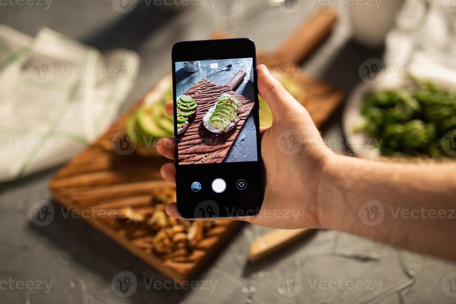 Hands take pictures on smartphone of two beautiful healthy sour cream and avocado sandwiches lying on board on the table. Social media and food concept photo