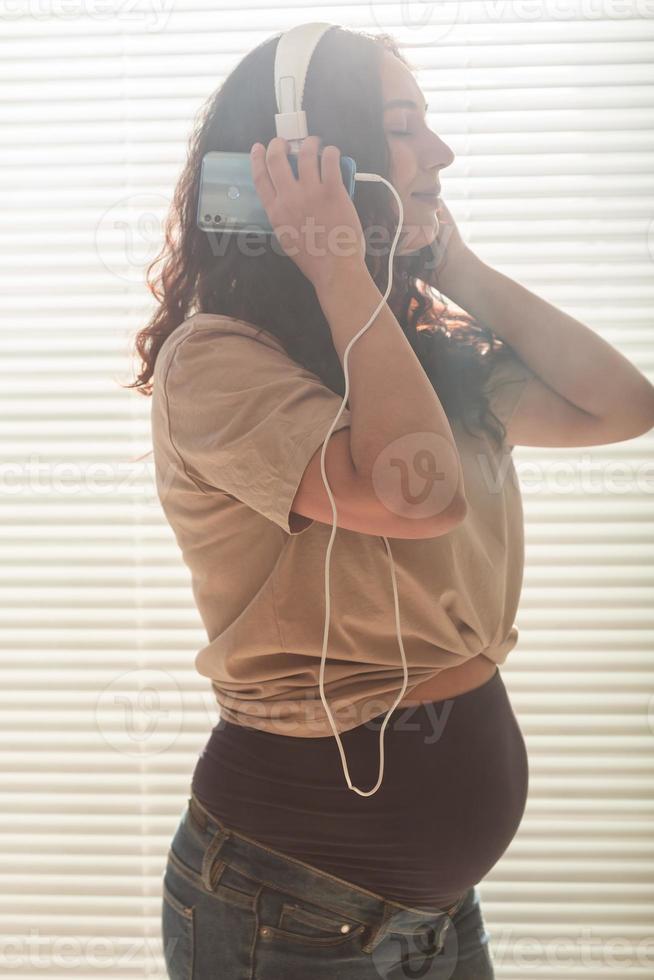 Curly-haired brunette pacified pregnant woman listens to music using smartphone and headphones. Concept of a soothing mood before meeting baby. photo