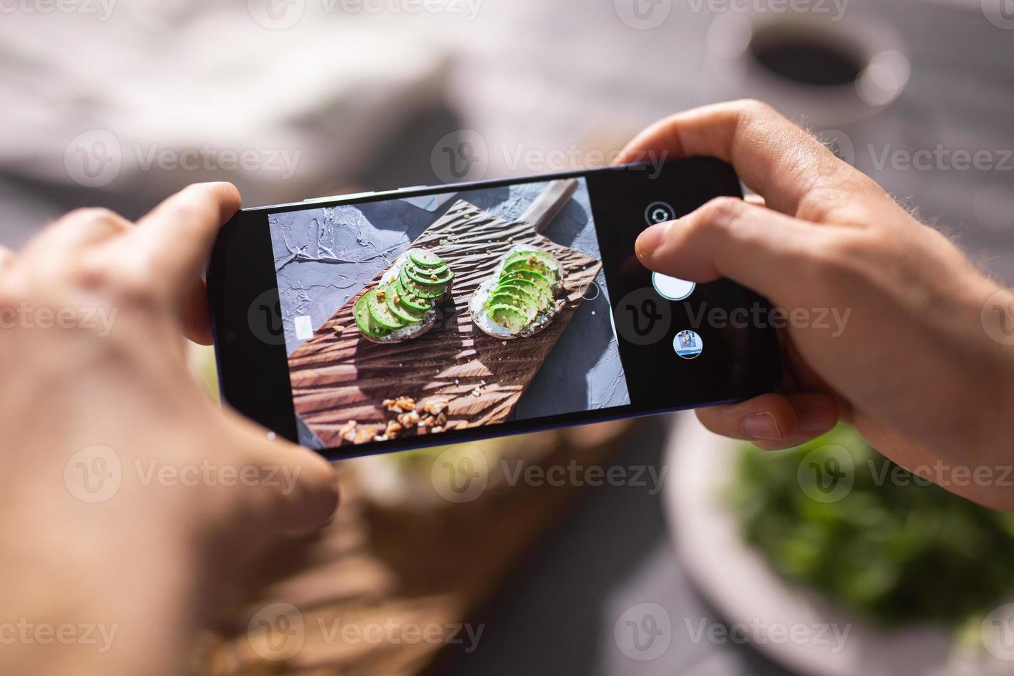Hands take pictures on smartphone of two beautiful healthy sour cream and avocado sandwiches lying on board on the table. Social media and food concept photo