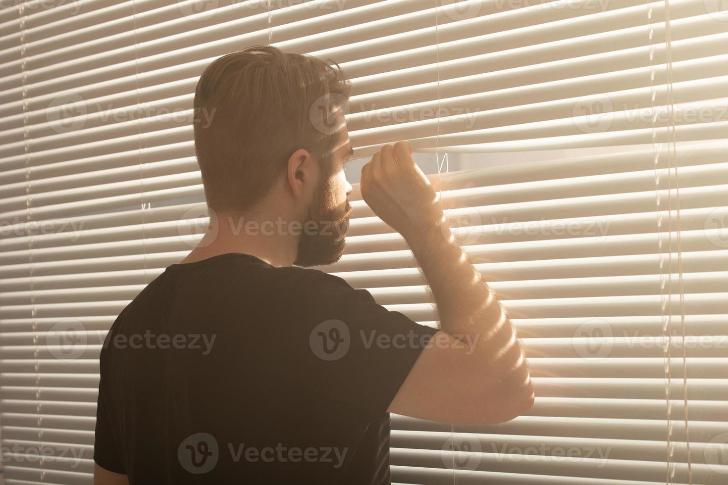 la vista trasera de un joven con barba se asoma por un agujero en las persianas de la ventana y mira hacia la calle. concepto de vigilancia y curiosidad foto