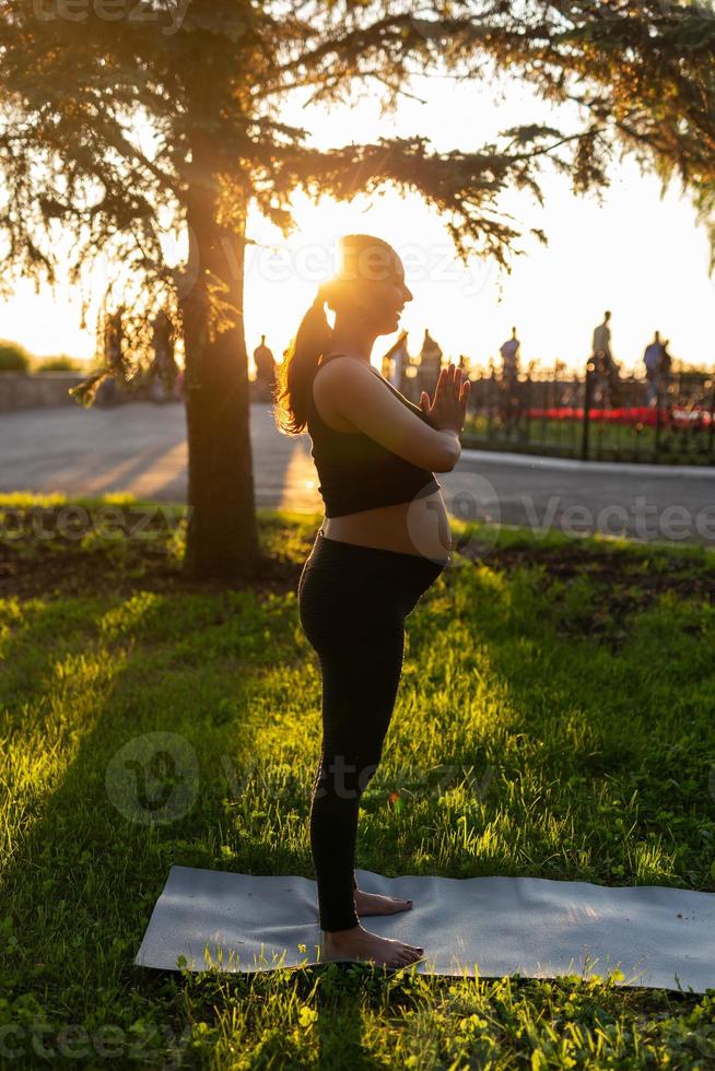 Side view of young woman expecting baby in black suit stands on rug barefoot and meditates on warm sunny summer day. Concept of sports and meditation activities photo