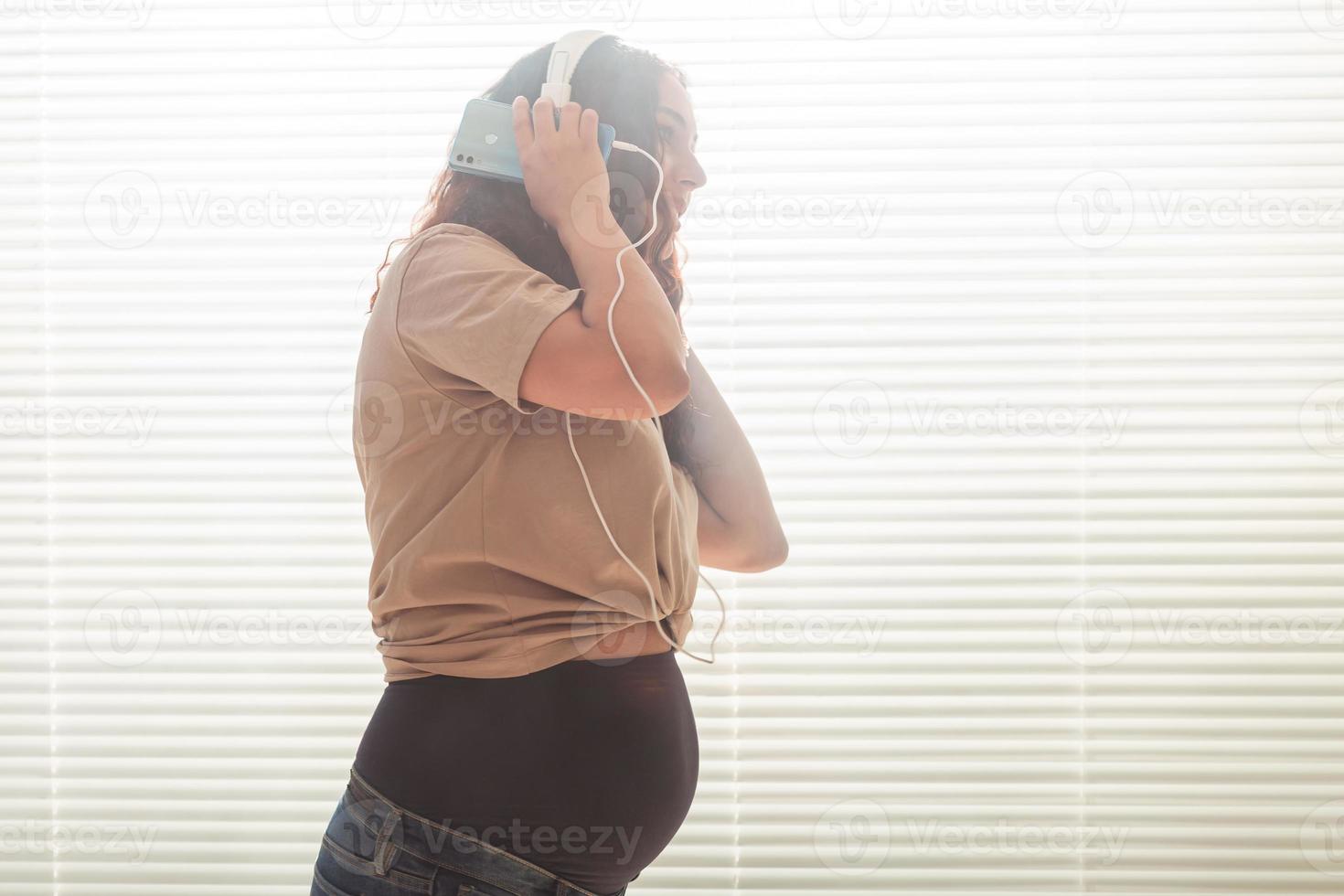 Curly-haired brunette pacified pregnant woman listens to pleasant classical music using smartphone and headphones. Concept of a soothing mood before meeting baby. Copyspace photo