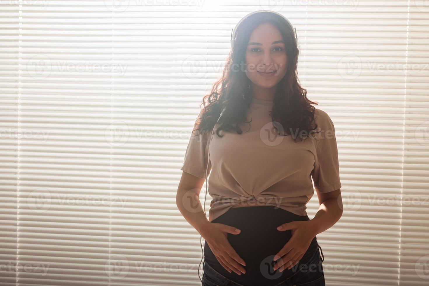 Curly-haired brunette pacified pregnant woman listens to pleasant classical music using smartphone and headphones. Concept of a soothing mood before meeting baby. Copyspace photo