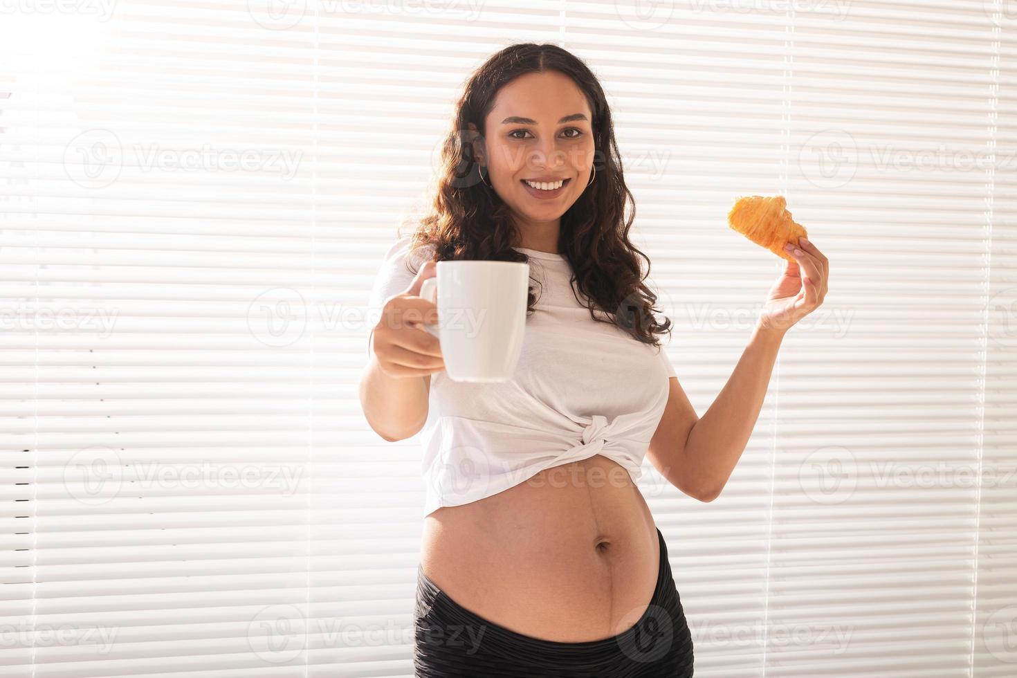 Beautiful pregnant woman holding croissant and cup of coffee in her hands during morning breakfast. Concept of good health and positive attitude while expecting baby photo
