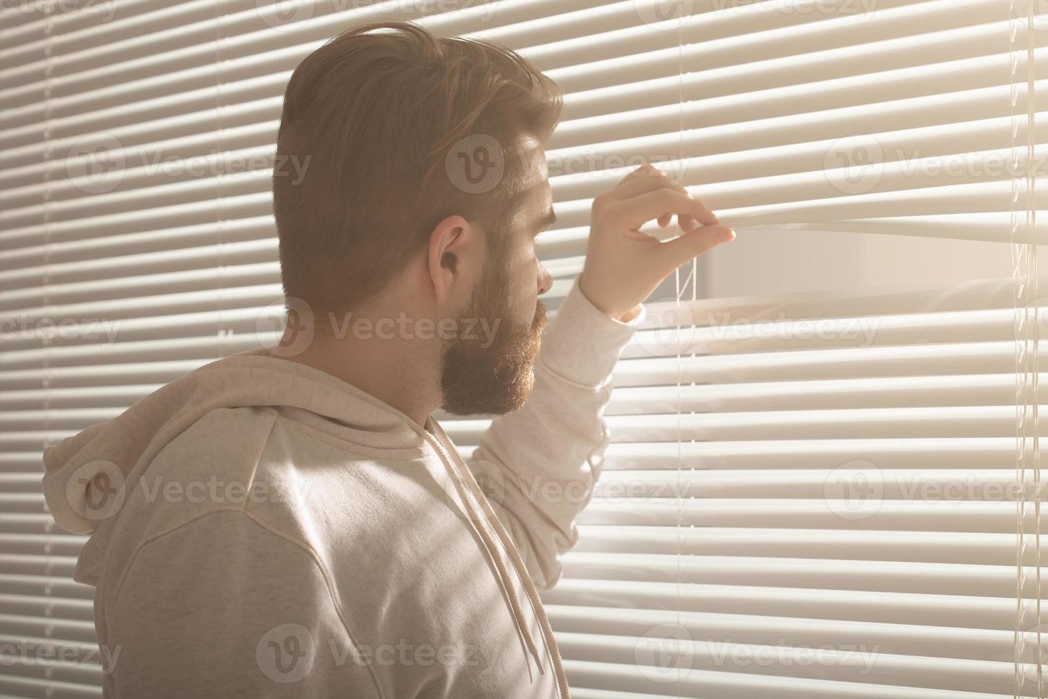 Rear view of young man with beard peeks through hole in the window blinds and looks out into the street. Surveillance and curiosity concept photo