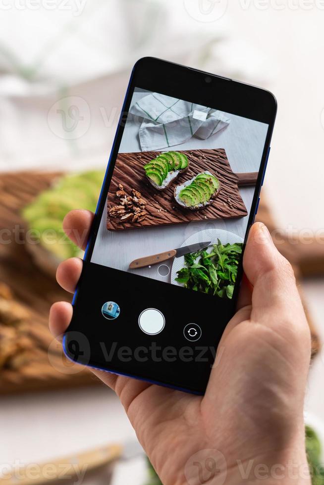 Hands take pictures on smartphone of two beautiful healthy sour cream and avocado sandwiches lying on board on the table. Social media and food concept photo