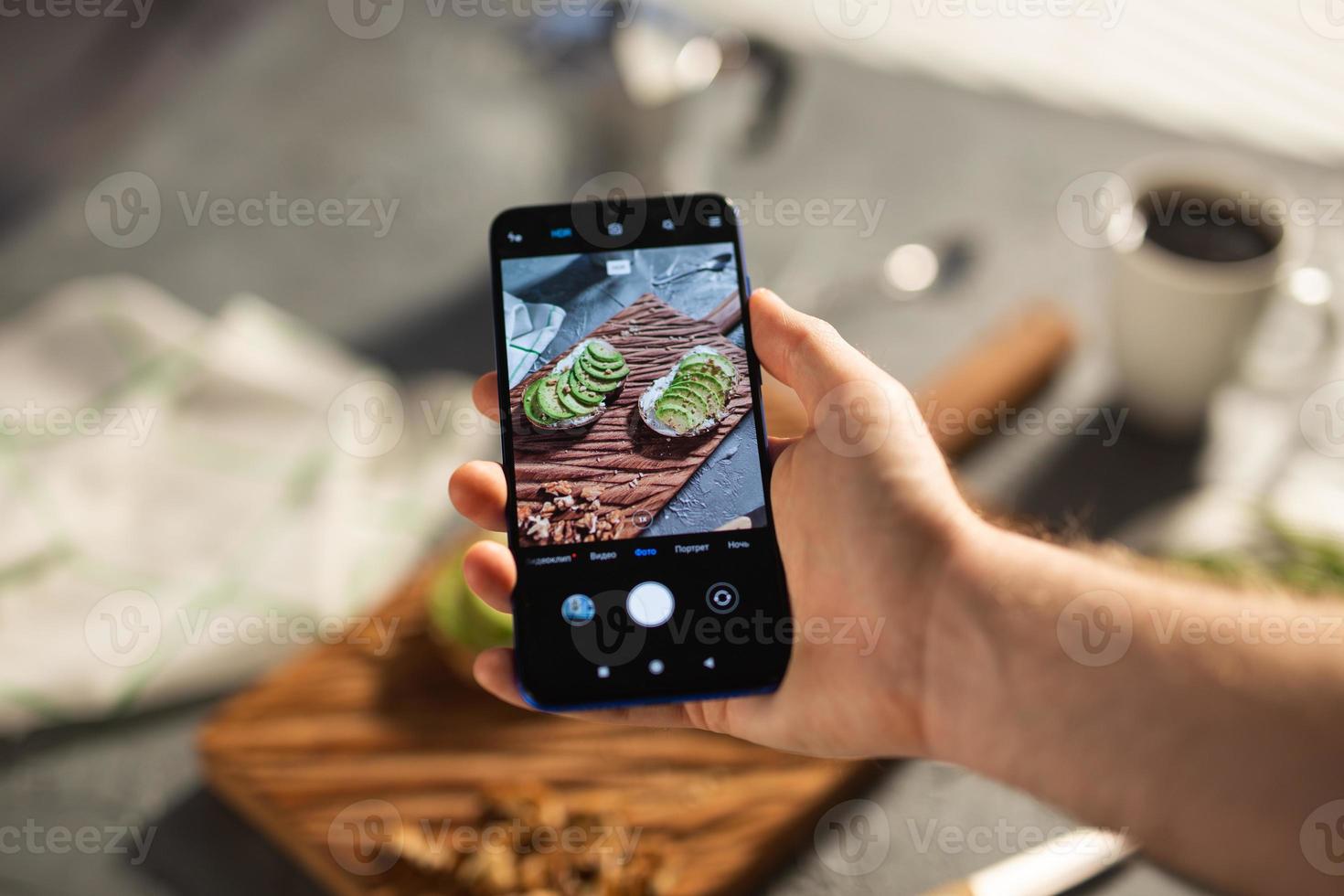 Hands take pictures on smartphone of two beautiful healthy sour cream and avocado sandwiches lying on board on the table. Social media and food concept photo