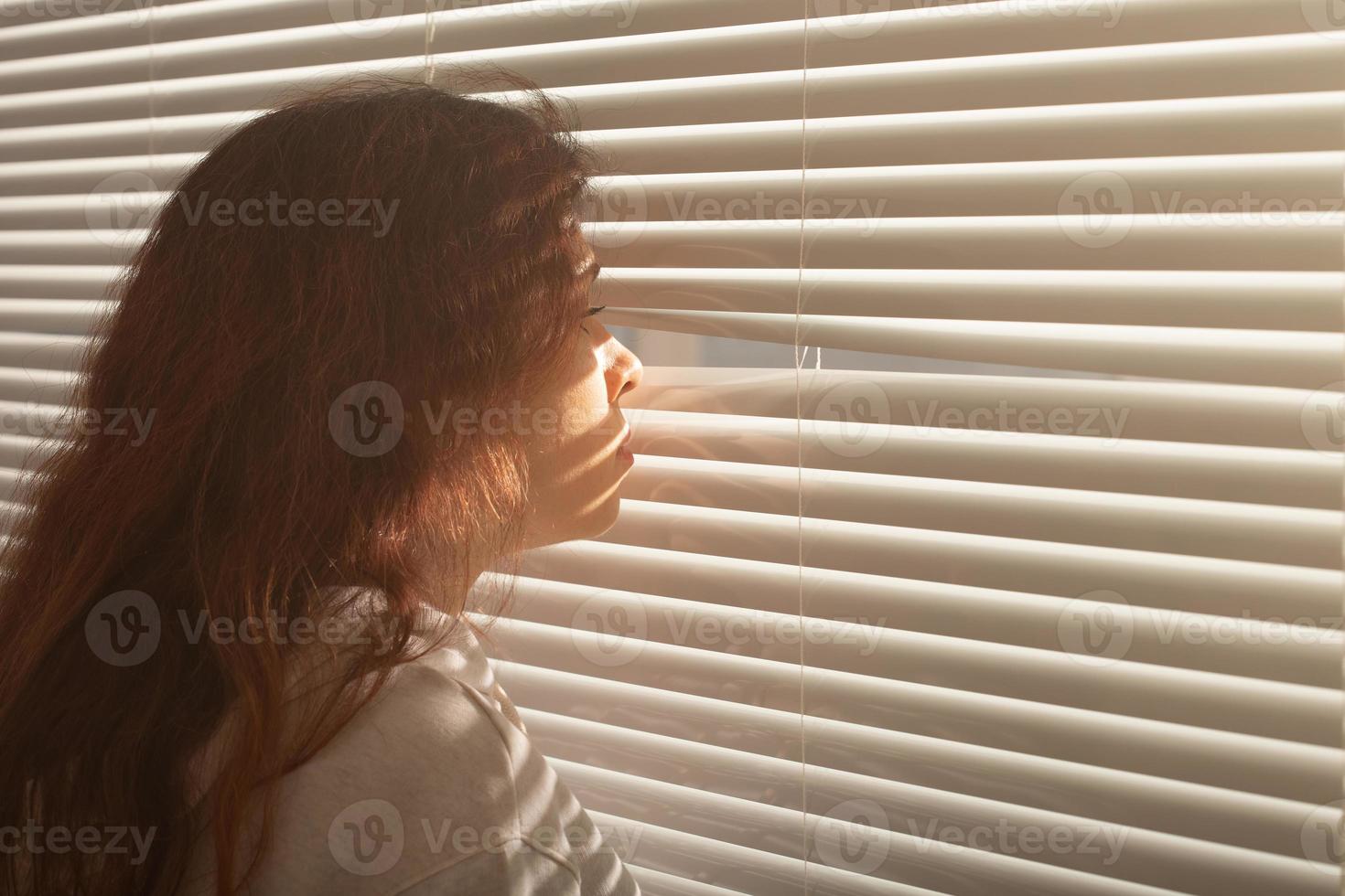 la vista trasera de una hermosa joven con el pelo largo se asoma por un agujero en las persianas de la ventana y mira por la ventana. concepto de vigilancia y curiosidad foto