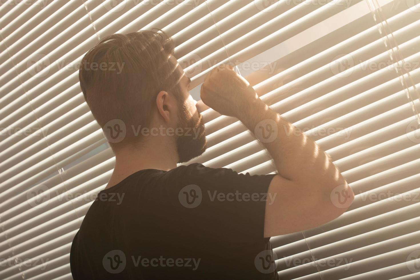 Rear view of young man with beard peeks through hole in the window blinds and looks out into the street. Surveillance and curiosity concept photo