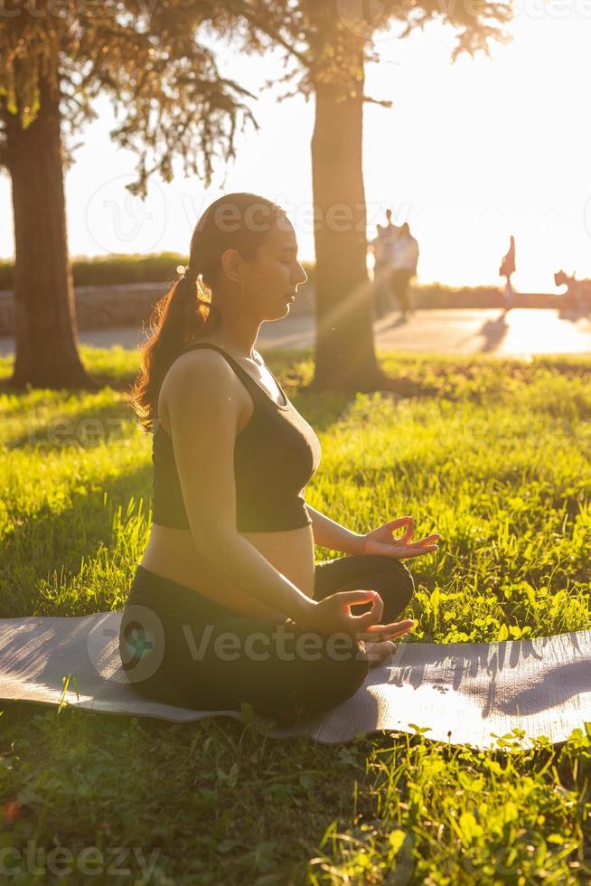 Cute young caucasian pregnant woman is meditating while sitting on a rug on the lawn on a sunny summer evening. Concept of pacification and energy boost photo