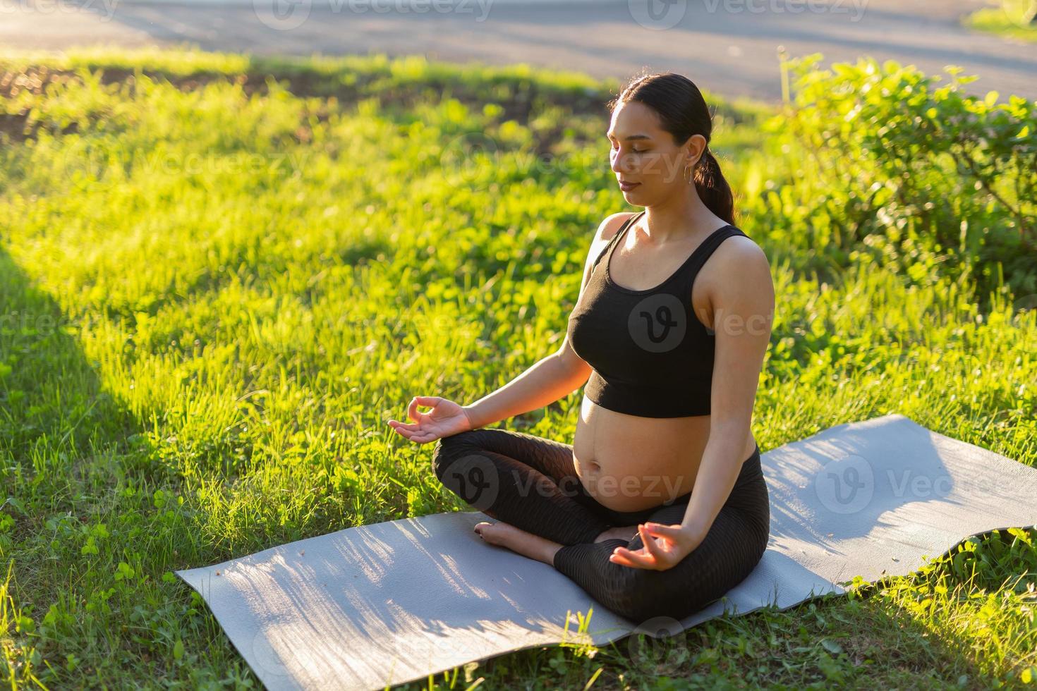 Una joven y pacífica mujer embarazada positiva con traje de gimnasia hace yoga y medita sentada en una alfombra sobre hierba verde en un día soleado y cálido de verano. concepto de preparación para el parto y actitud positiva foto