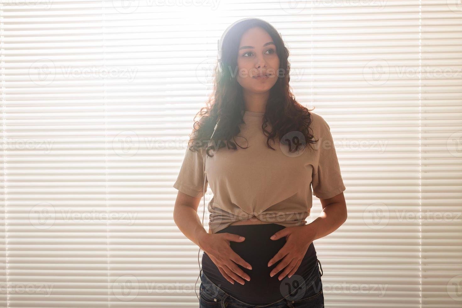 Curly-haired brunette pacified pregnant woman listens to pleasant classical music using smartphone and headphones. Concept of a soothing mood before meeting baby. Copyspace photo