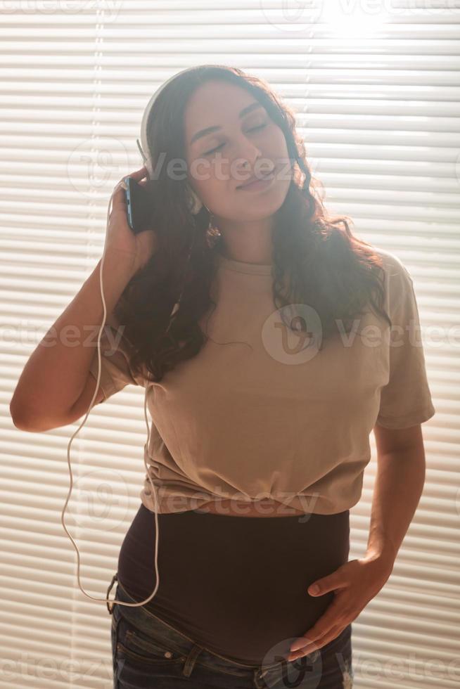 Curly-haired brunette pacified pregnant woman listens to pleasant music using smartphone and headphones. Concept of a soothing mood before meeting baby. photo