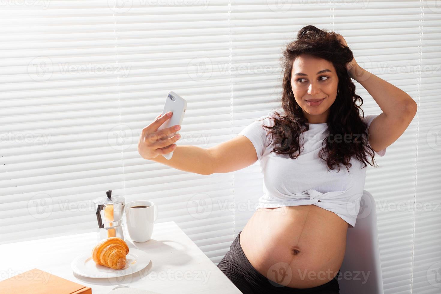 Pregnant woman takes selfie on her smartphone while breakfast. Maternity leave and technologies concept. photo