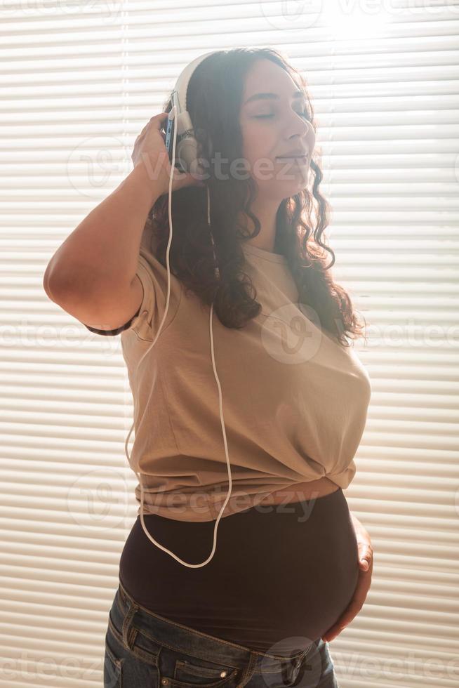 Curly-haired brunette pacified pregnant woman listens to pleasant classical music using smartphone and headphones. Concept of a soothing mood before meeting baby. photo